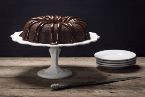 A chocolate bundt cake with ganache icing sits atop a cake stand on a wooden table.